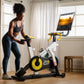 Woman in black sports bra and blue leggings using white and yellow stationary bike with mounted tablet display in bright room