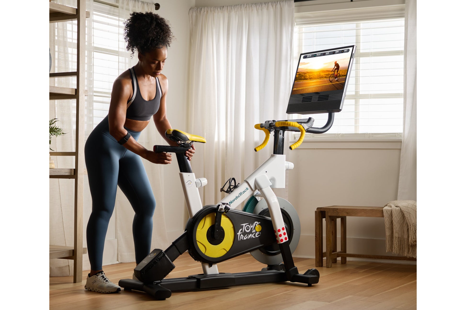 Woman in black sports bra and blue leggings using white and yellow stationary bike with mounted tablet display in bright room