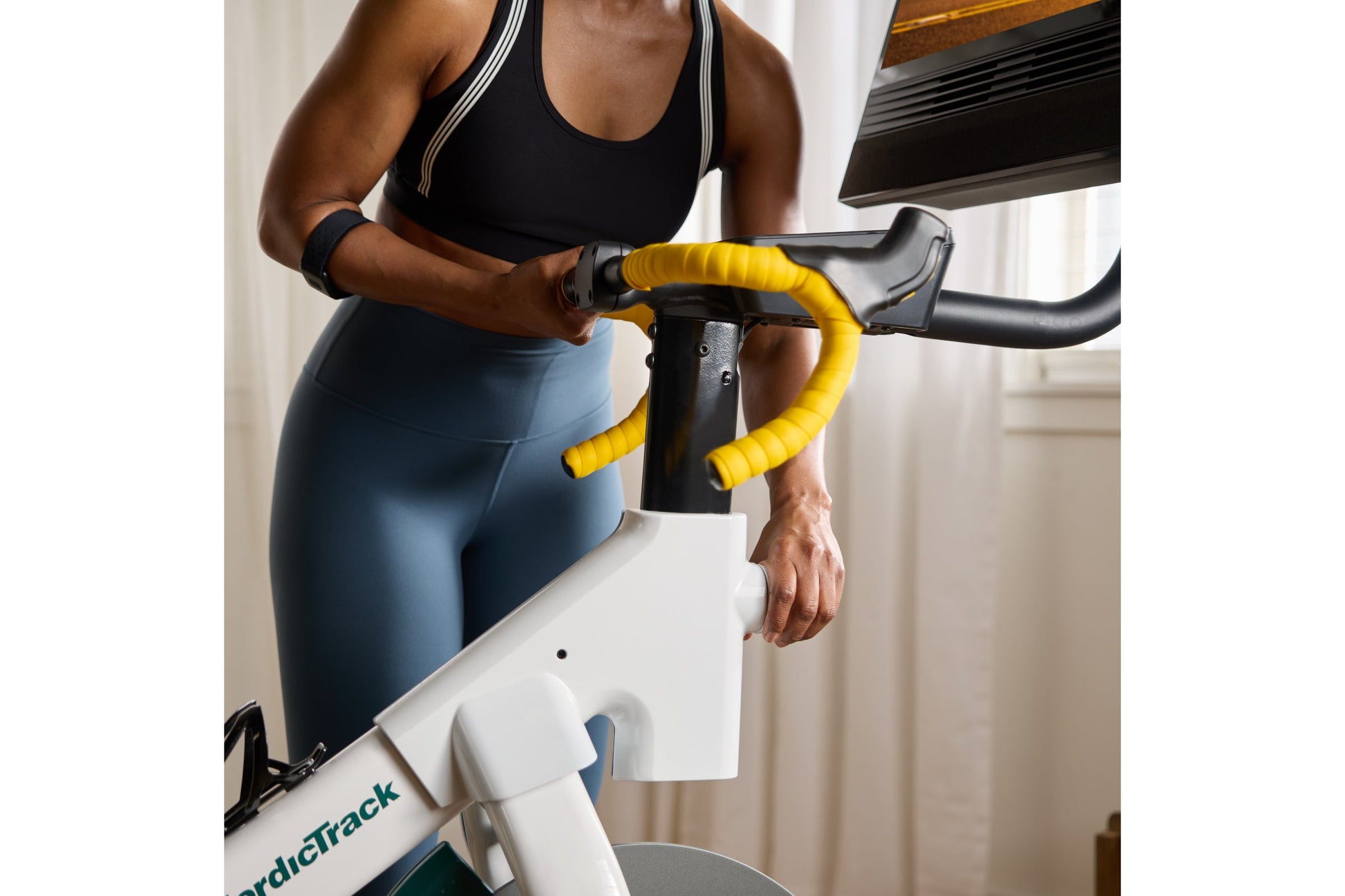 Woman in black sports bra and blue leggings riding stationary bike with yellow handlebar tape, white and teal frame
