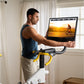 Man in blue tank top and beige shorts riding white and yellow indoor stationary bike with digital display showing cycling vid