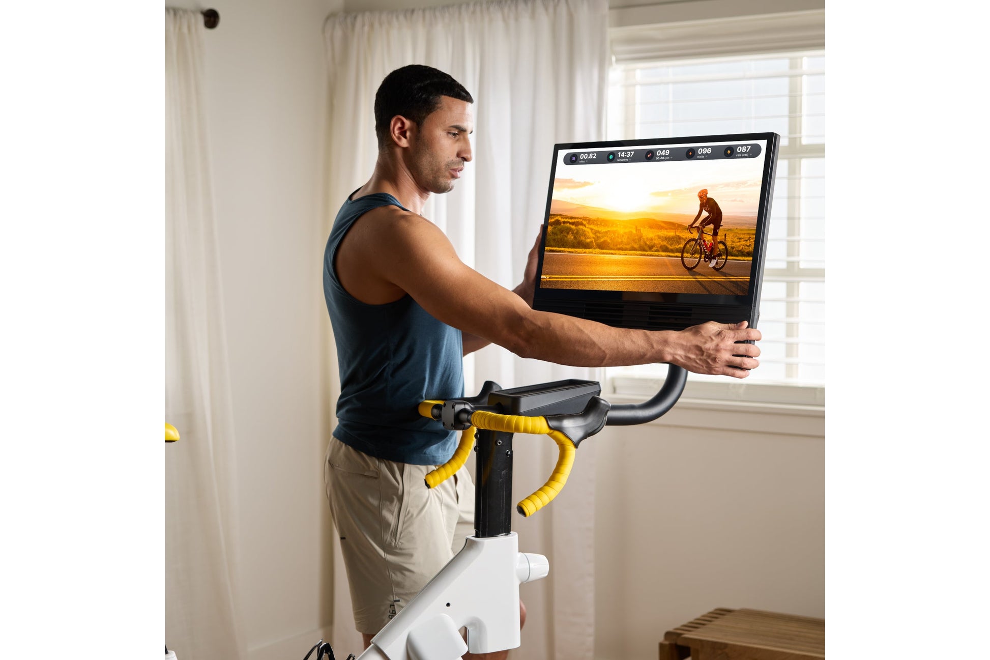 Man in blue tank top and beige shorts riding white and yellow indoor stationary bike with digital display showing cycling vid