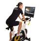 Woman wearing black cycling jersey with "CITY" text riding stationary bike with yellow accents and digital screen displaying 