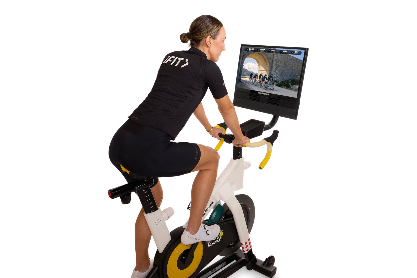 Woman wearing black cycling jersey with "CITY" text riding stationary bike with yellow accents and digital screen displaying 