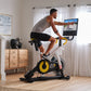 Man on stationary indoor bike with yellow and black wheel, gray tank top, black shorts, holding yellow handlebars while viewi