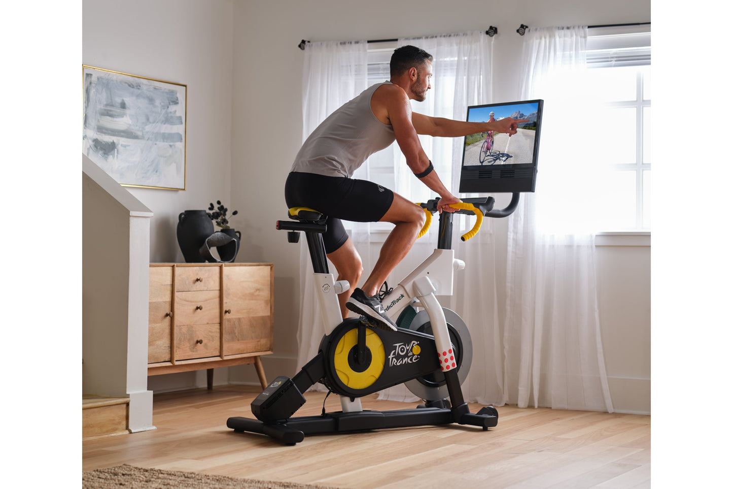 Man on stationary indoor bike with yellow and black wheel, gray tank top, black shorts, holding yellow handlebars while viewi