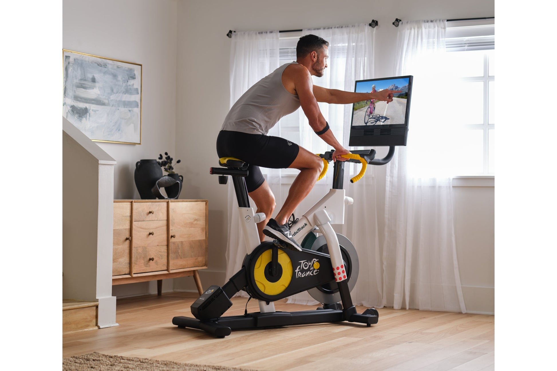 Man on stationary indoor bike with yellow and black wheel, gray tank top, black shorts, holding yellow handlebars while viewi