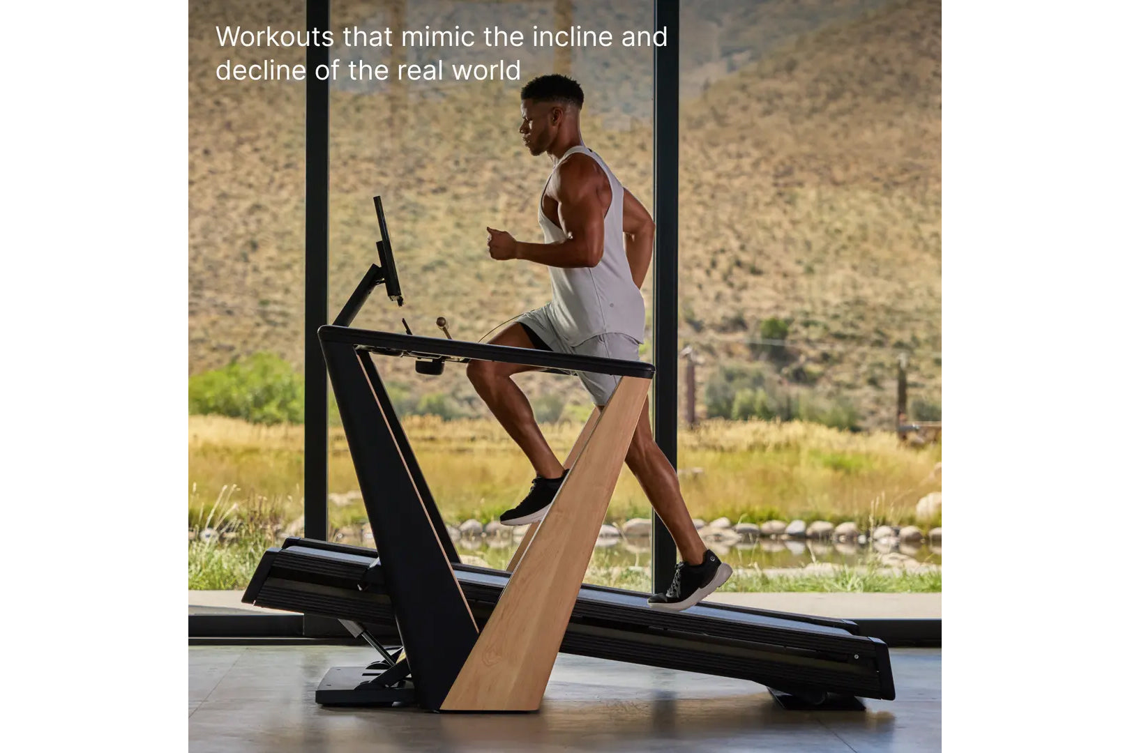 Man in white tank top running on black and wood NordicTrack treadmill with incline feature, facing large windows overlooking 