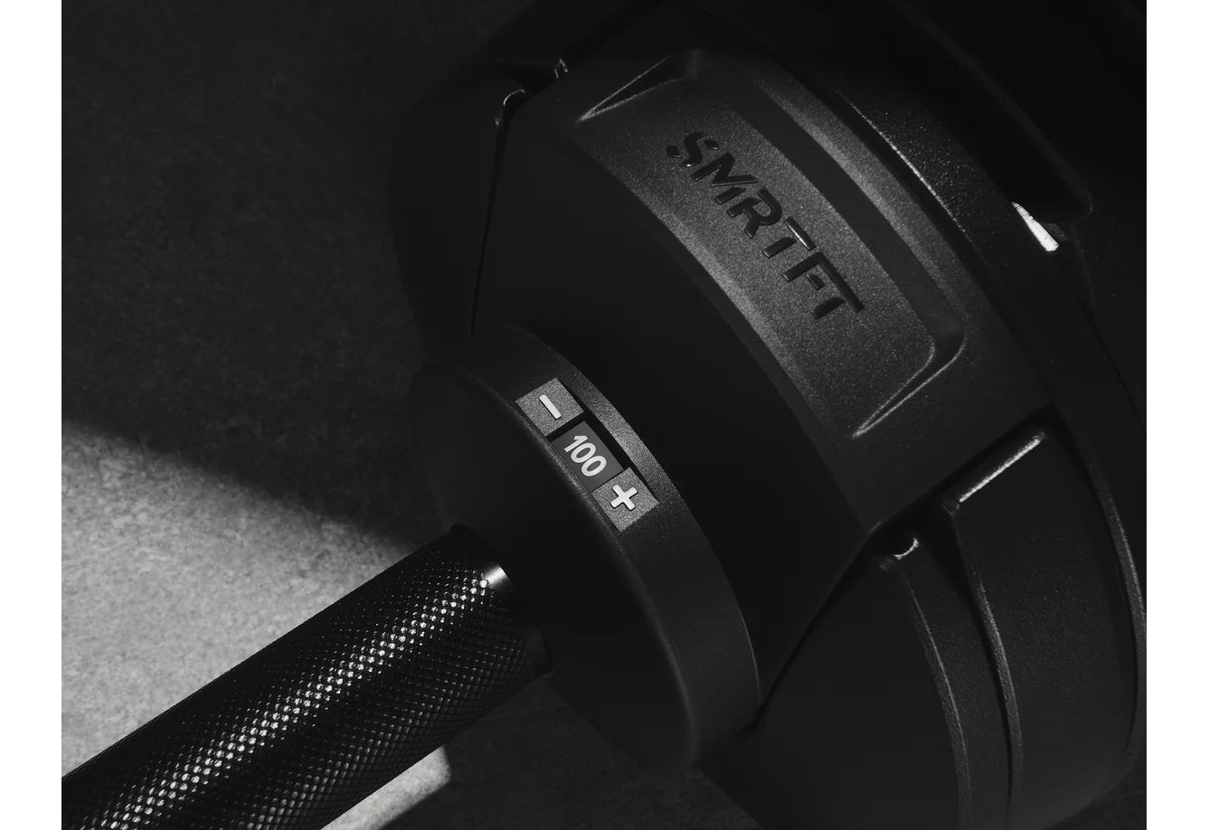 Black adjustable dumbbell marked "NÜOBELL" and "100+" shown in flat lay on textured surface