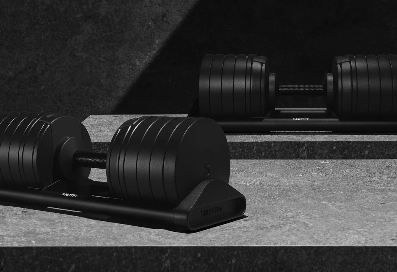 Matte black adjustable dumbbells with ribbed weight plates on stand, displayed on concrete floor