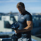 Man wearing slate blue NÜO t-shirt holding black adjustable dumbbell on rooftop with city skyline background