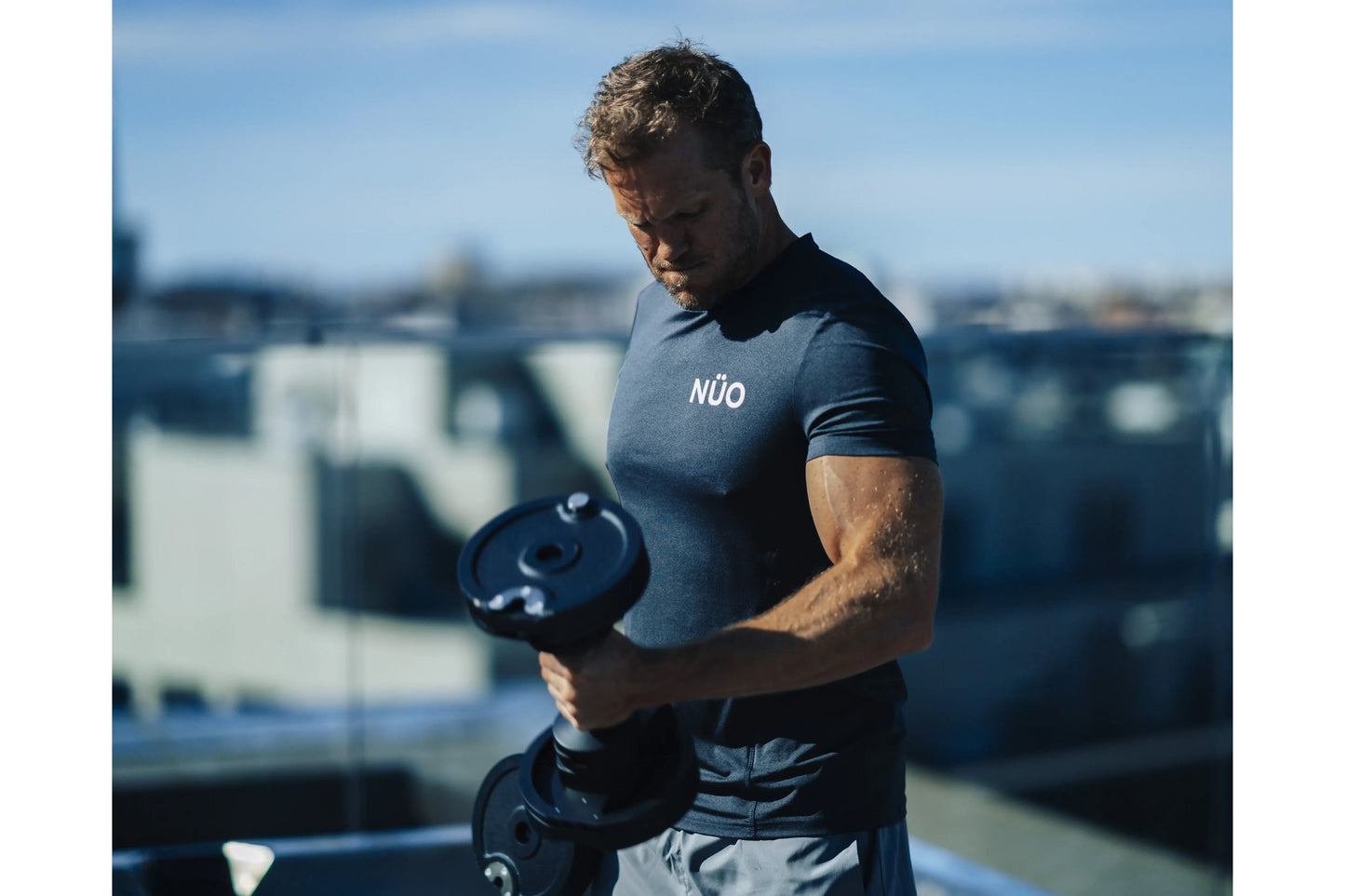 Man wearing slate blue NÜO t-shirt holding black adjustable dumbbell on rooftop with city skyline background