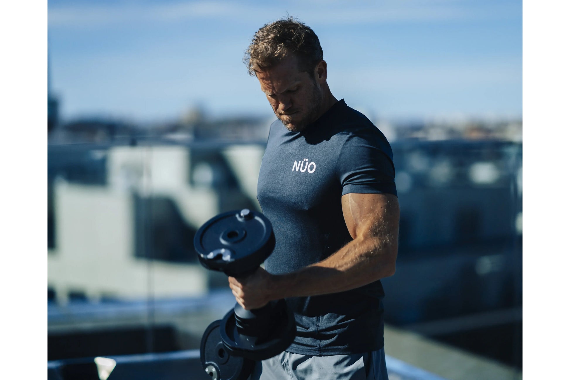 Man wearing slate blue NÜO t-shirt holding black adjustable dumbbell on rooftop with city skyline background