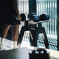 Person holding black adjustable dumbbell during workout with matching dumbbells on stand, white socks visible, indoor gym set