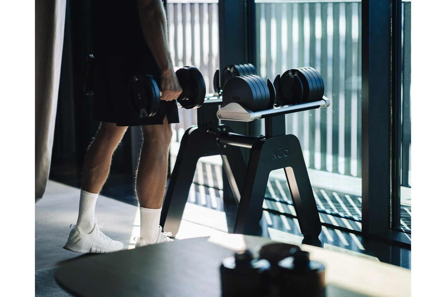 Person holding black adjustable dumbbell during workout with matching dumbbells on stand, white socks visible, indoor gym set