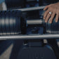 Adjustable dumbbell with black weight plates and textured gray handle, held by hand wearing silver ring on metal rack