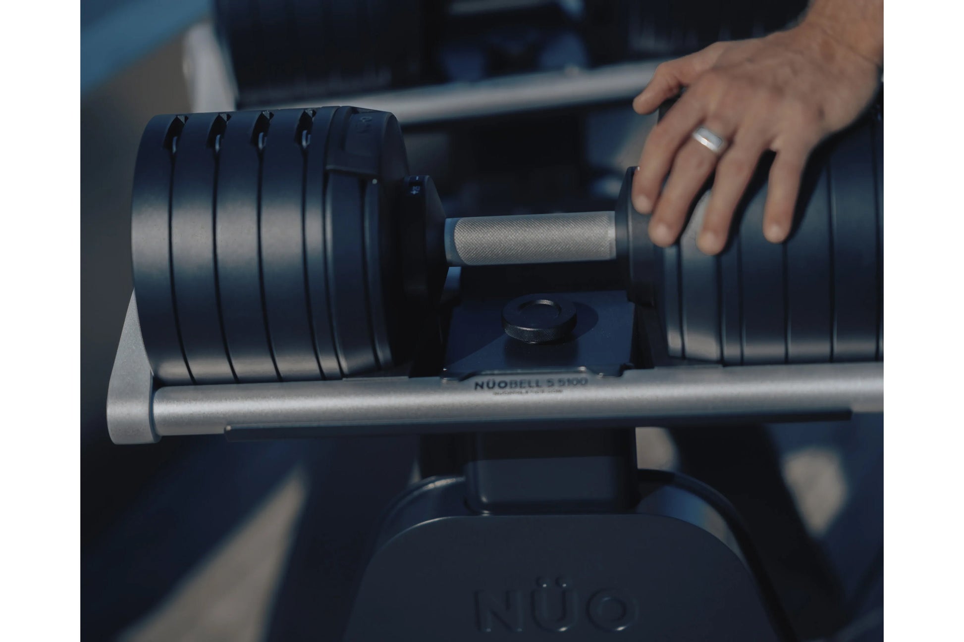 Adjustable dumbbell with black weight plates and textured gray handle, held by hand wearing silver ring on metal rack