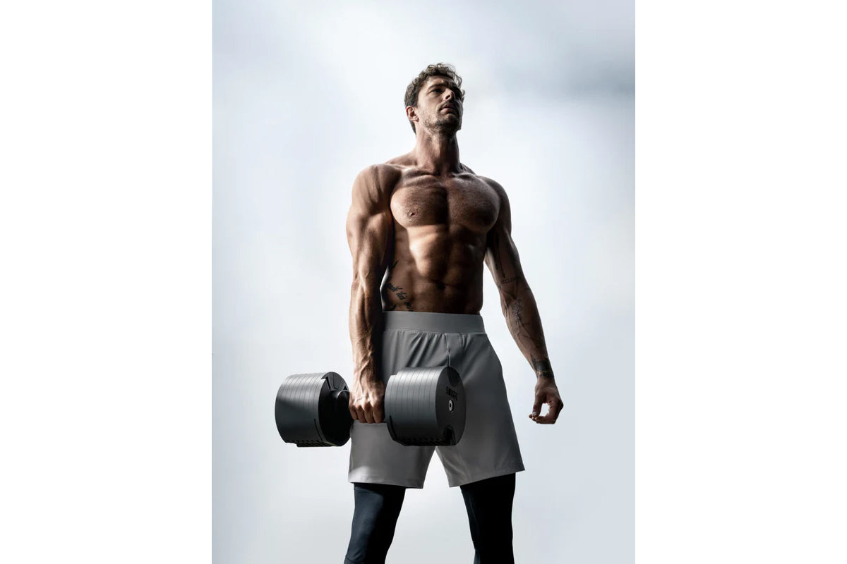 Shirtless man holding black adjustable dumbbells wearing gray shorts against white background