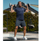 Man wearing navy t-shirt and charcoal shorts holding pair of adjustable dumbbells overhead outdoors