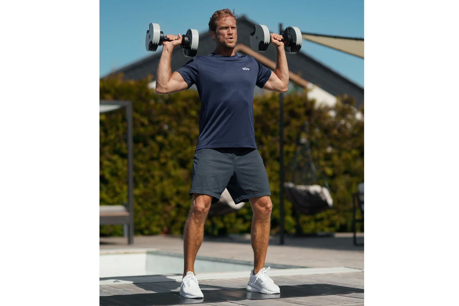 Man wearing navy t-shirt and charcoal shorts holding pair of adjustable dumbbells overhead outdoors