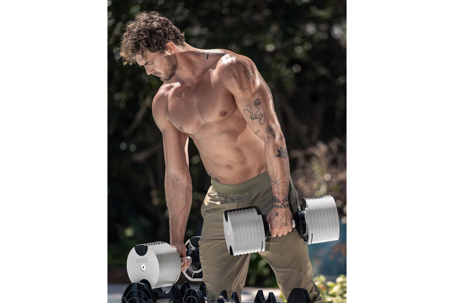 Man with curly hair and tattoos lifting white and black NÜOBELL adjustable dumbbells outdoors, wearing olive shorts