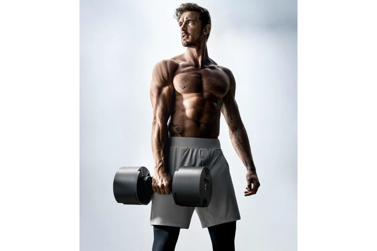 Muscular man wearing gray shorts holding matte black adjustable dumbbells, shown against white background