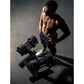 Shirtless man holding adjustable dumbbell while standing next to dumbbell rack on dark floor, wearing navy shorts and white s