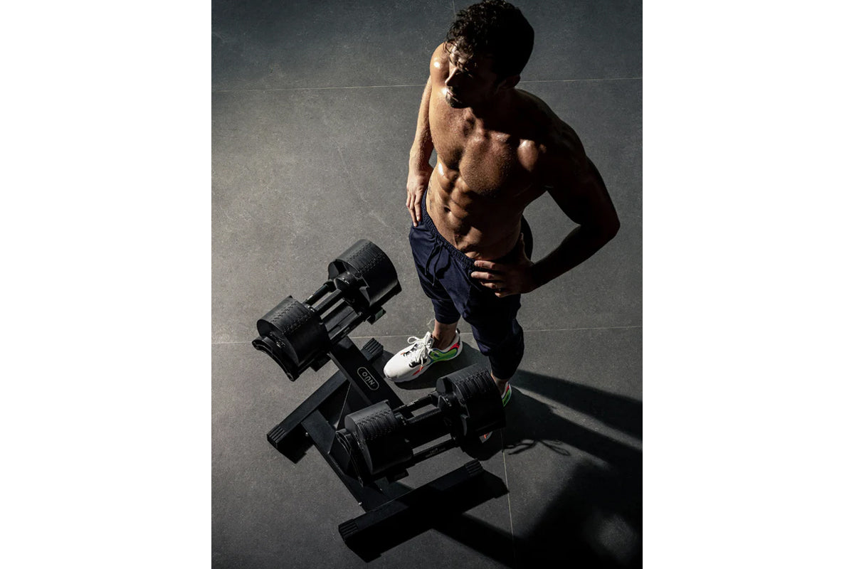 Shirtless man holding adjustable dumbbell while standing next to dumbbell rack on dark floor, wearing navy shorts and white s
