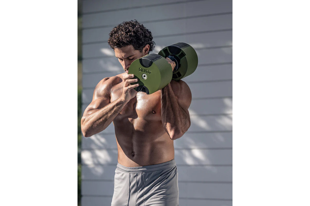 Man performing shoulder press with olive green adjustable dumbbell, shirtless, outdoors against gray wall