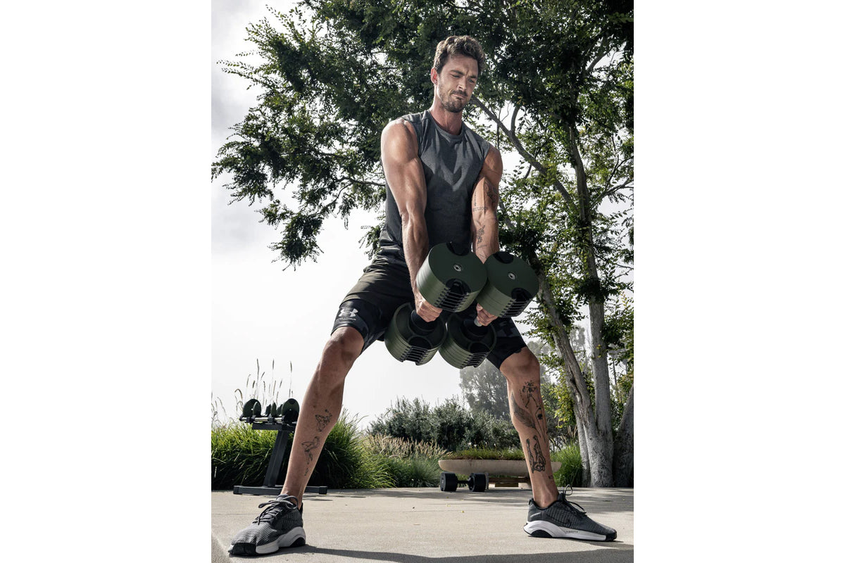 Man holding olive and black adjustable dumbbells in wide stance during outdoor workout session