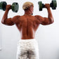 Shirtless man holding green and black adjustable dumbbells overhead, viewed from behind, wearing white patterned shorts