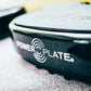 Black and white Power Plate vibration machine with circular logo, shown at angle on textured surface with tools blurred in ba