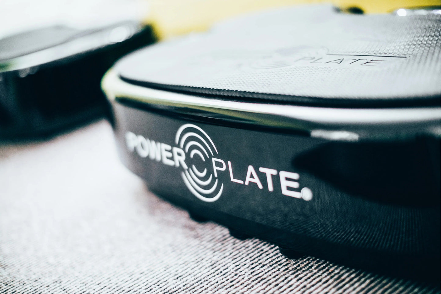 Black and white Power Plate vibration machine with circular logo, shown at angle on textured surface with tools blurred in ba
