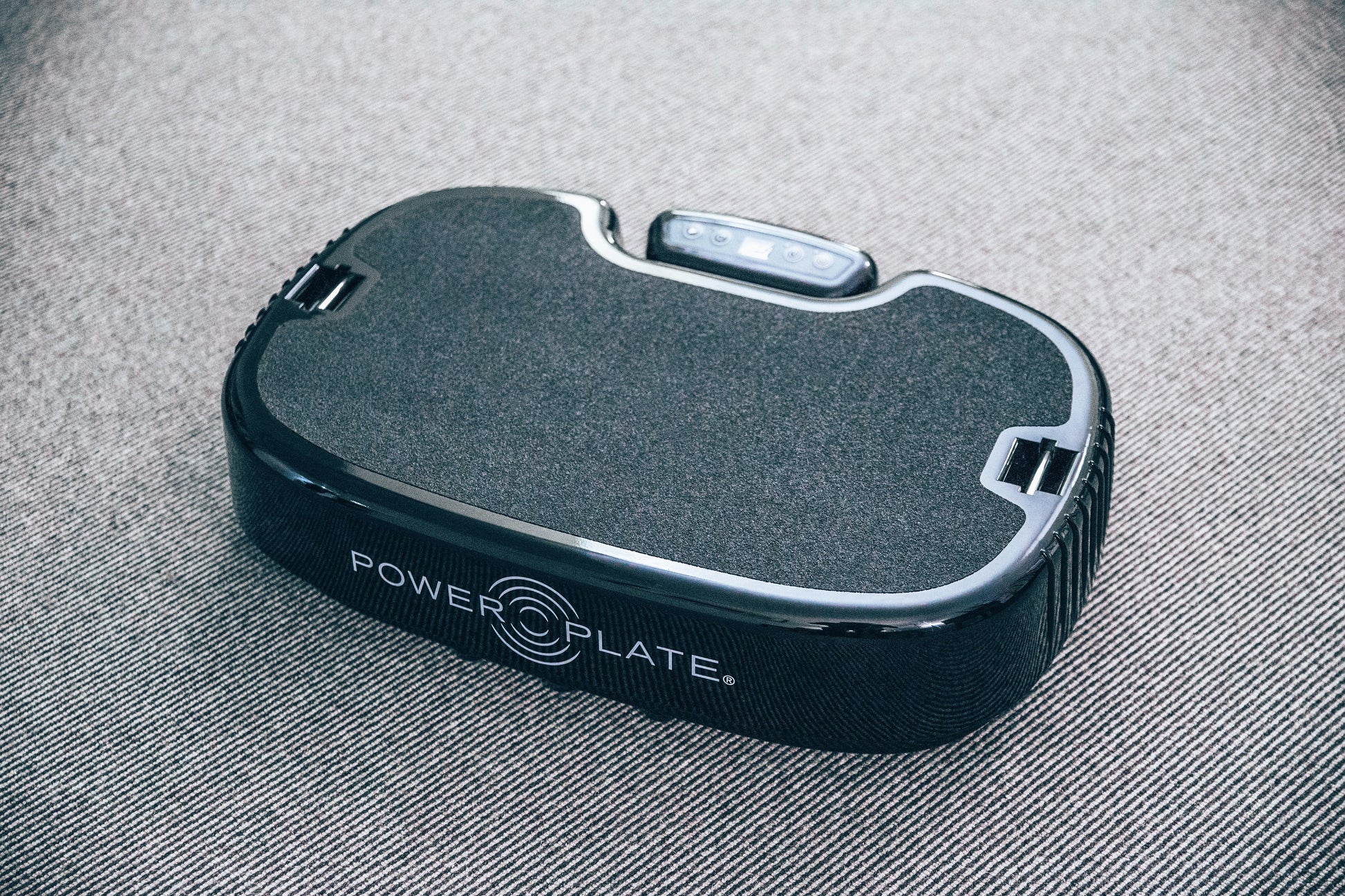 Power Plate Personal Vibration Machine — angled view showing textured non-slip surface, chrome trim, and control buttons on gray fabric background