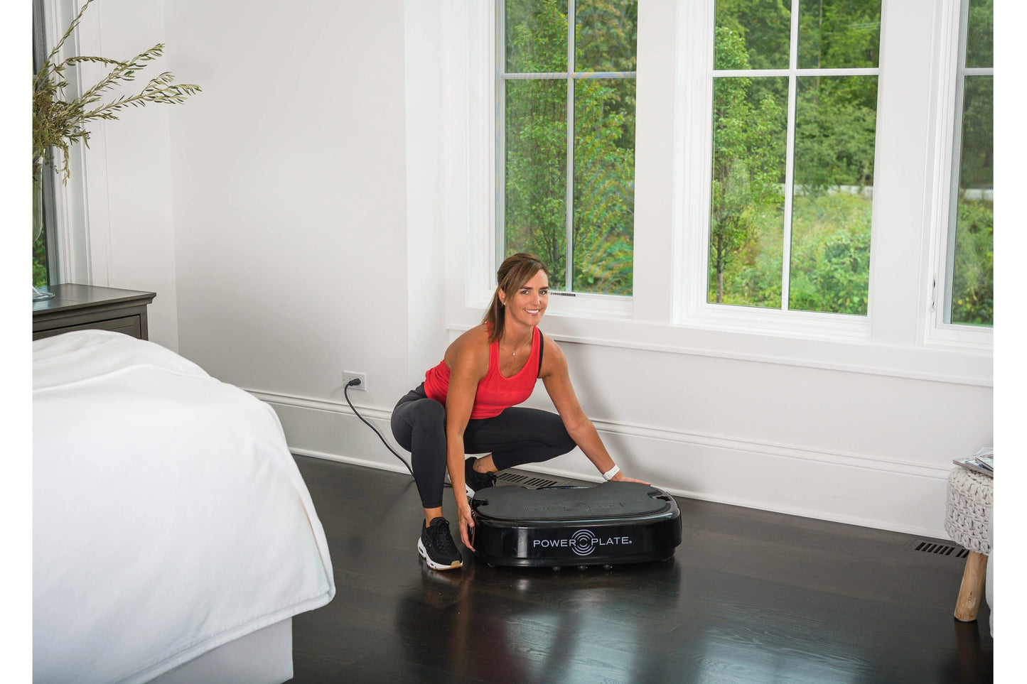 Woman in red tank top and black leggings using black Power Plate vibration machine in bedroom, worn by model