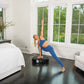 Woman in blue ribbed sports bra and leggings using black Power Plate vibration machine in bright bedroom with white bed and w