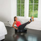 Woman in red tank top and black leggings using black Power Plate vibration machine on bedroom floor, legs extended