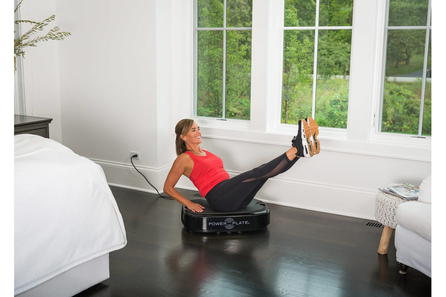 Woman in red tank top and black leggings using black Power Plate vibration machine on bedroom floor, legs extended