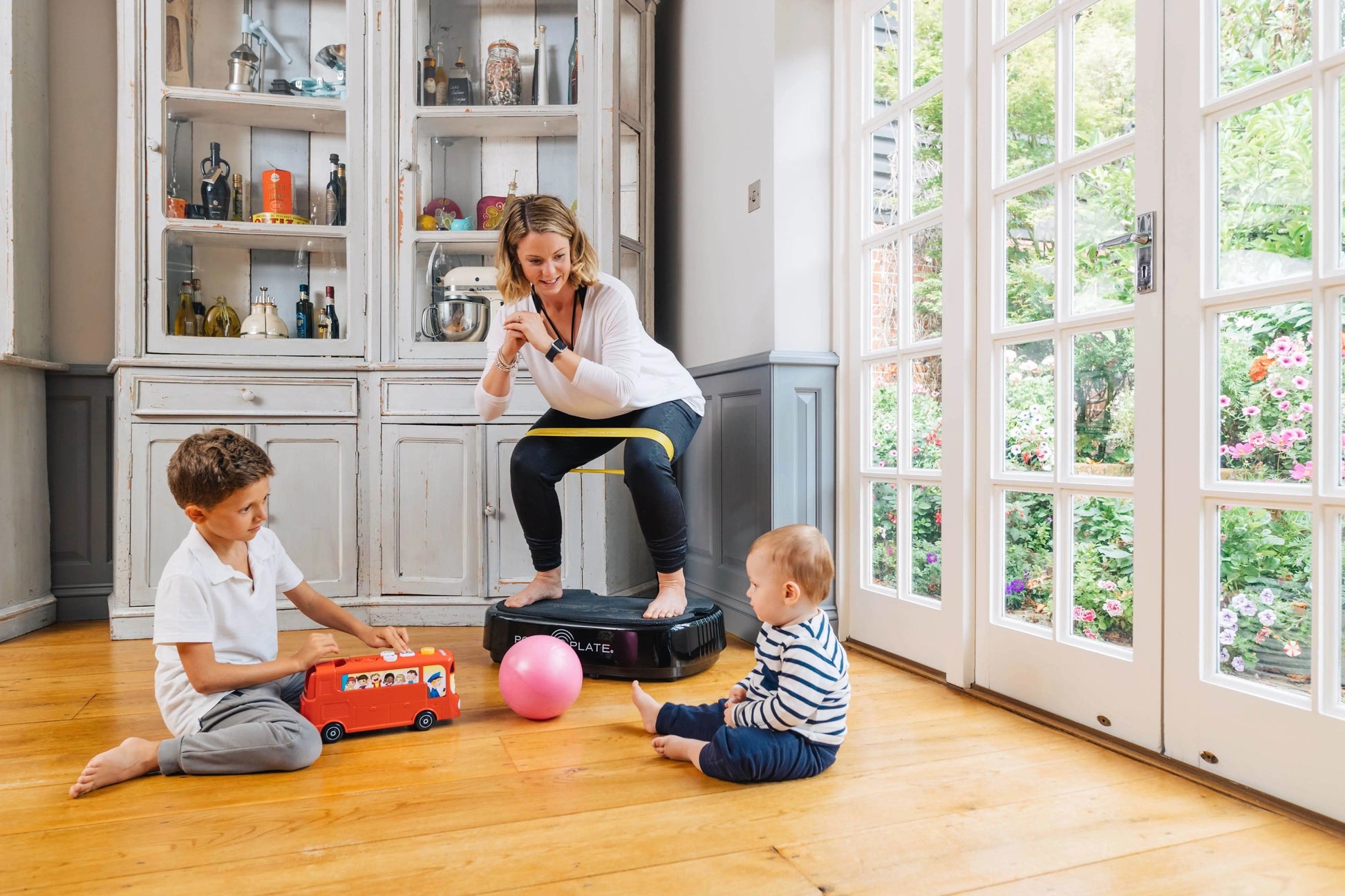 Woman in white top and black leggings with yellow waistband using black Power Plate vibration machine while two young childre