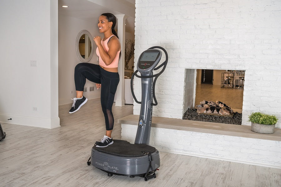 Woman using black Power Plate My7 vibration trainer in home gym, wearing pink sports bra and black leggings, reflected in mir