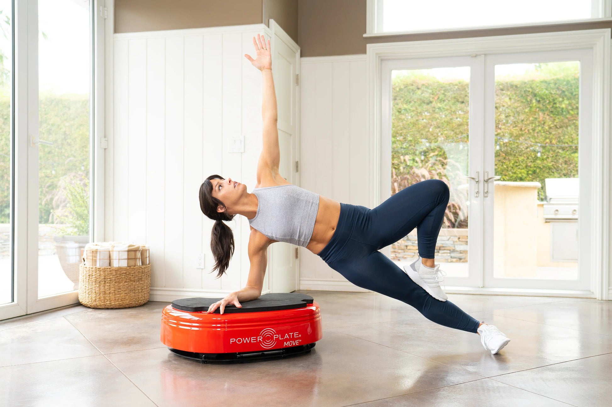 Woman in gray crop top and navy leggings performing a core exercise on a red Power Plate MOVE vibration trainer in a bright h