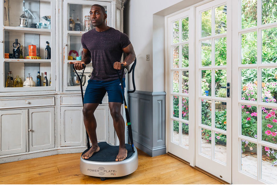Man wearing charcoal t-shirt and navy shorts standing on white Power Plate My5 vibration trainer in home gym setting