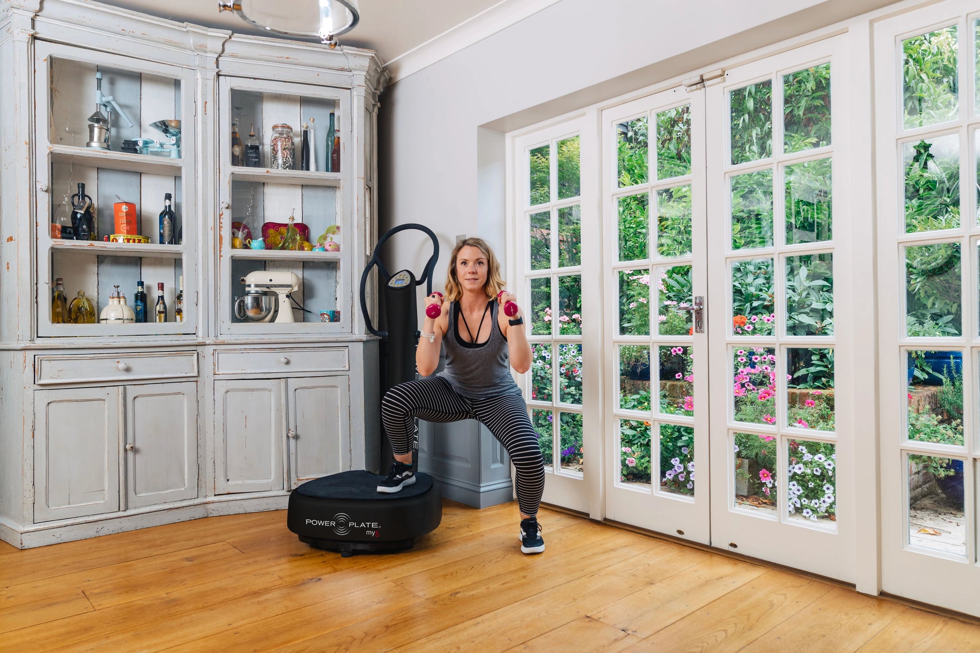 Woman exercising on black Power Plate My5 vibration trainer while holding pink dumbbells, wearing gray tank top and striped l