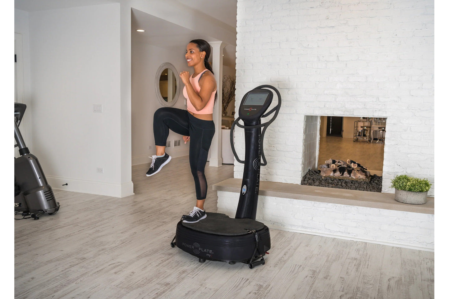 Woman in pink sports bra and black leggings using black Power Plate My7 vibration trainer in modern home gym with white brick