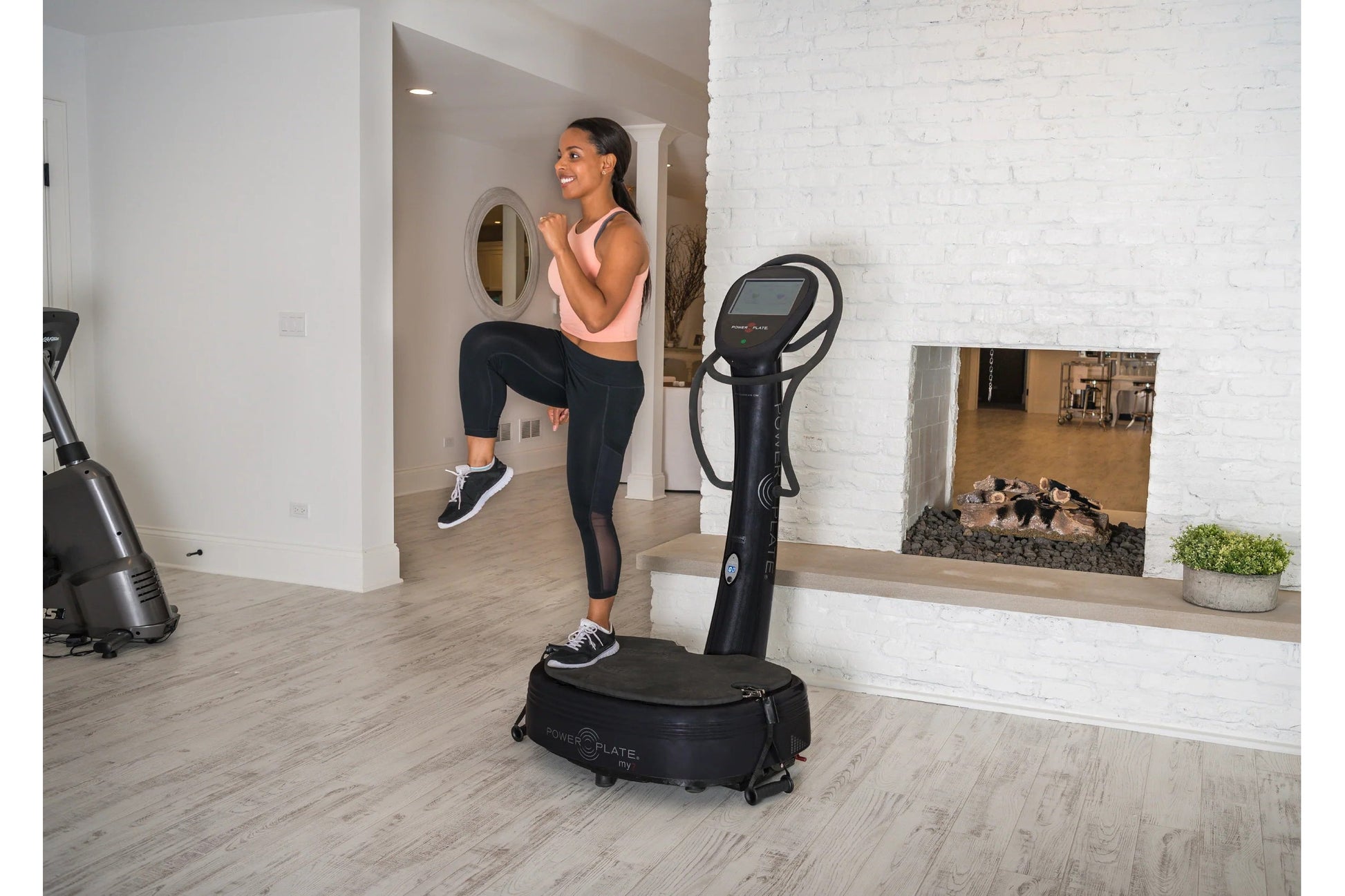 Woman in pink sports bra and black leggings using black Power Plate My7 vibration trainer in modern home gym with white brick