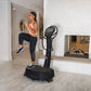 Woman in peach tank top and black leggings using black Power Plate vibration machine in modern studio setting with mirror ref