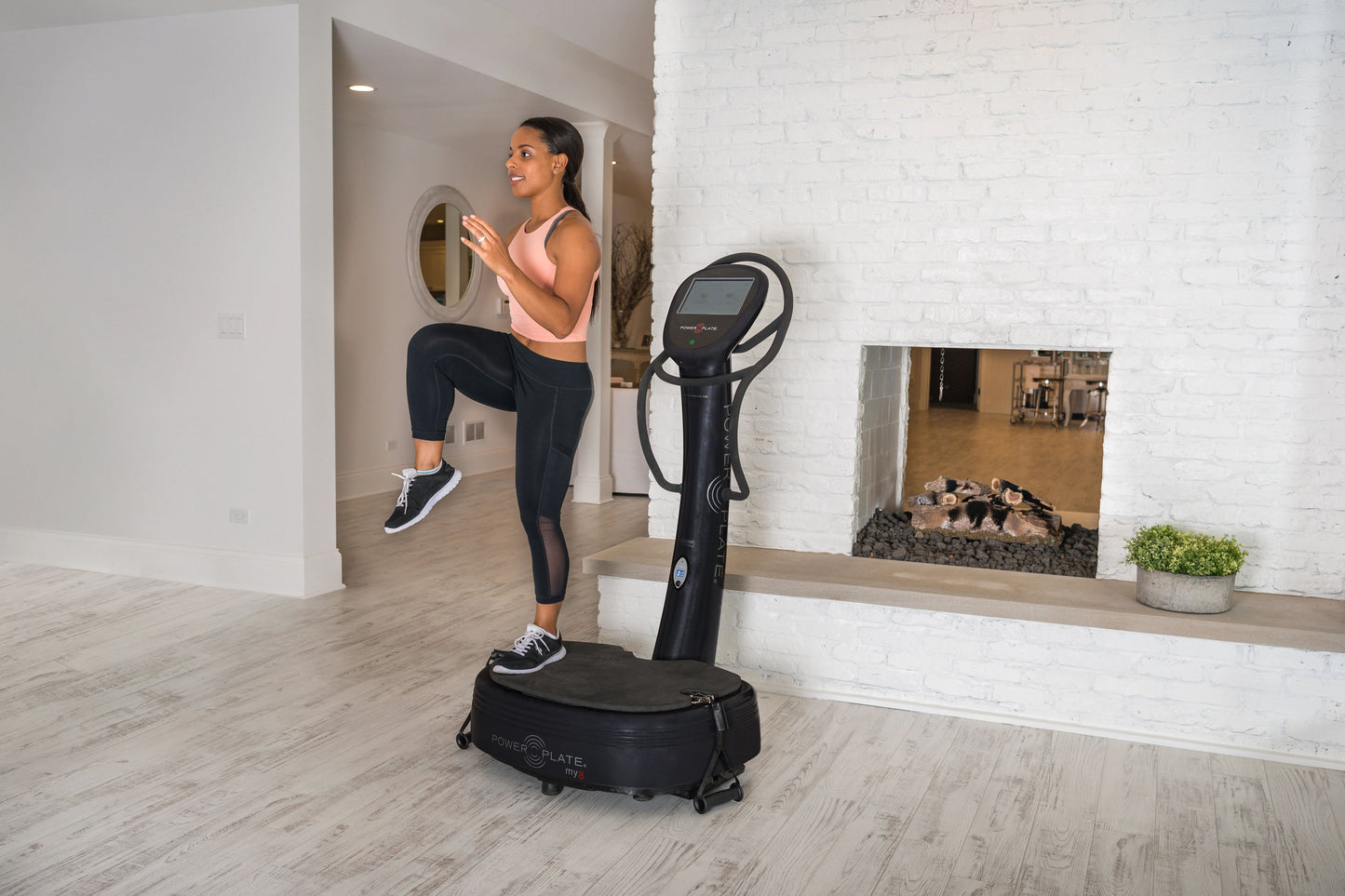Woman in peach tank top and black leggings using black Power Plate vibration machine in modern studio setting with mirror ref