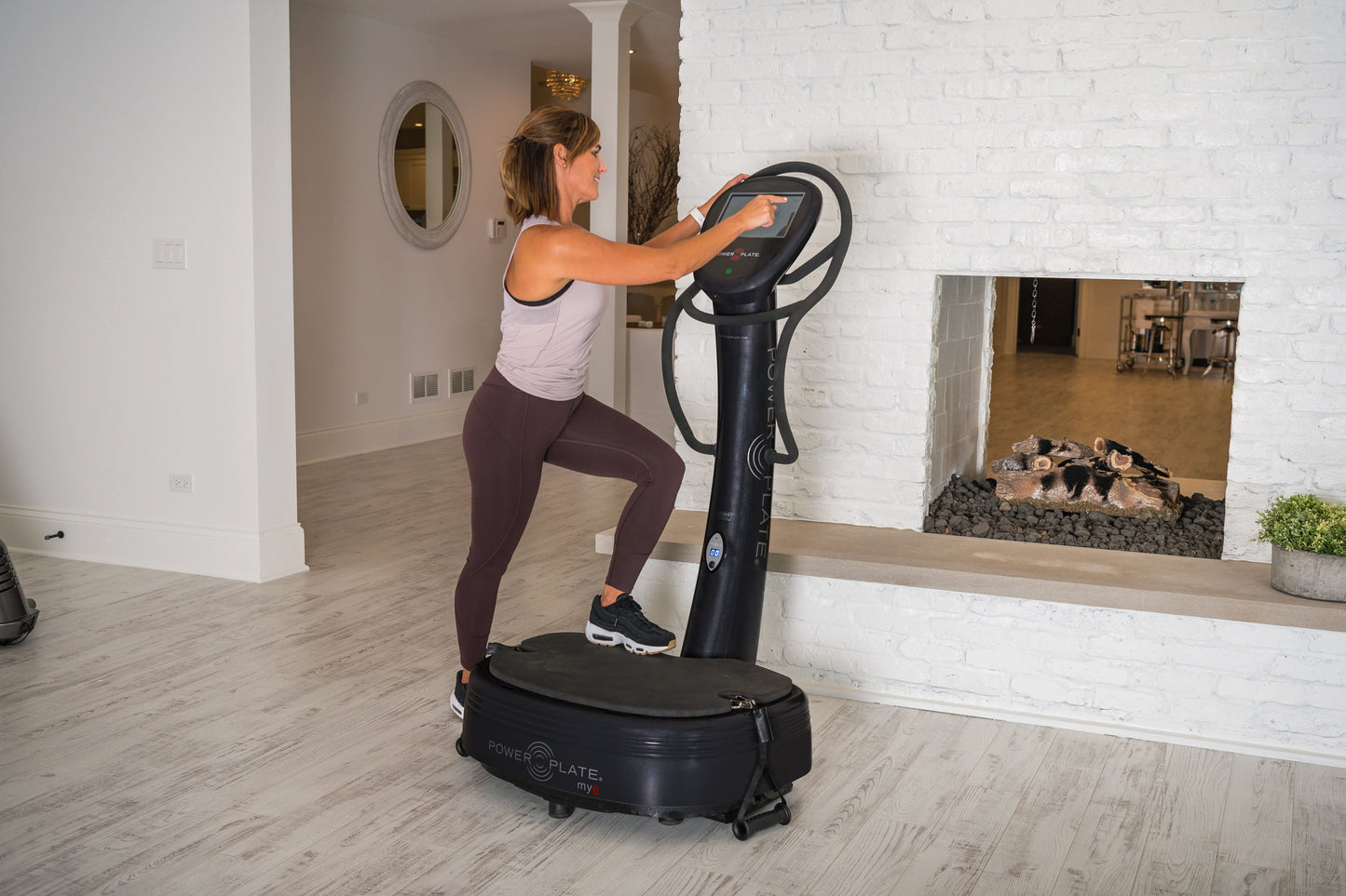 Woman in white tank top and burgundy leggings using black Power Plate My8 vibration trainer in modern gym setting