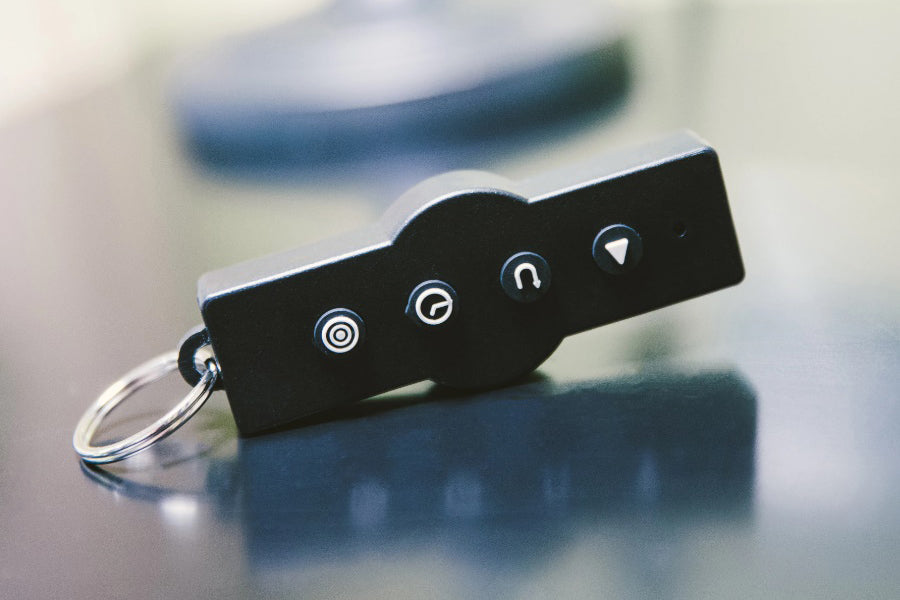 Black remote control with four silver icon buttons and metal keyring attached, on blurred background