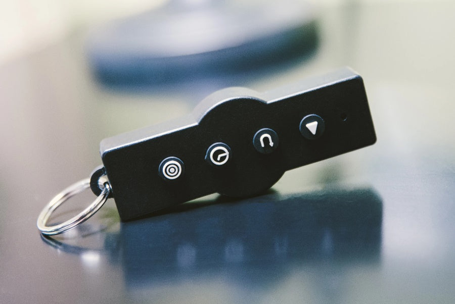 Black remote control with four white icon buttons and silver key ring, on neutral background