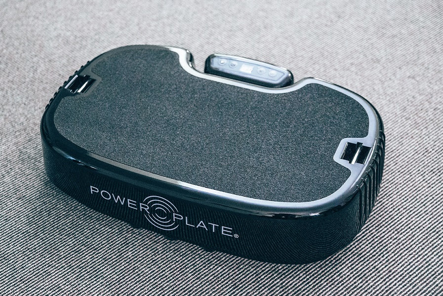 Black PowerPlate vibration machine with grey textured surface and control panel on top, flat lay view on grey background